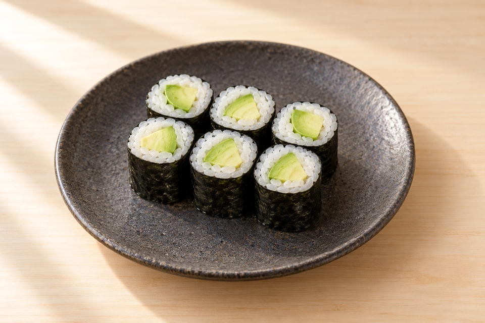 Six avocado maki pieces with a nori exterior and pale green filling on a charcoal ceramic plate. — AI recipe illustration